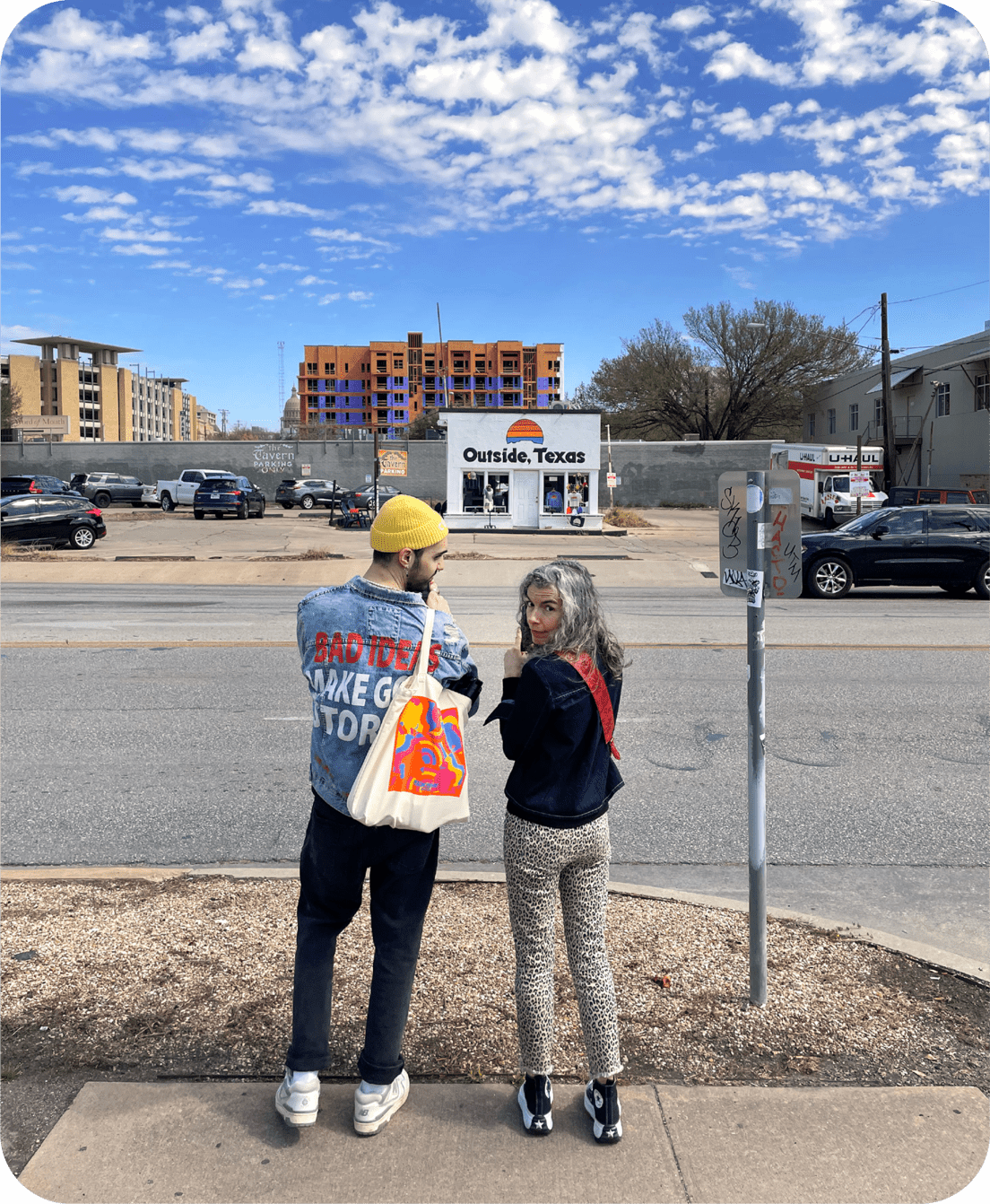 Nick Tetrault and Kristen Hughes are seen standing on a sidewalk in Austin, Texas, facing away from the camera, looking back over their shoulders. Nick is on the left, wearing a yellow beanie and a denim jacket with bold red text that reads "BAD IDEAS MAKE GOOD STORIES." Kristen, on the right, has curly gray hair and is wearing a dark jacket with leopard print pants. There is a small building in the background labeled "Outside, Texas," and a partly cloudy sky overhead.