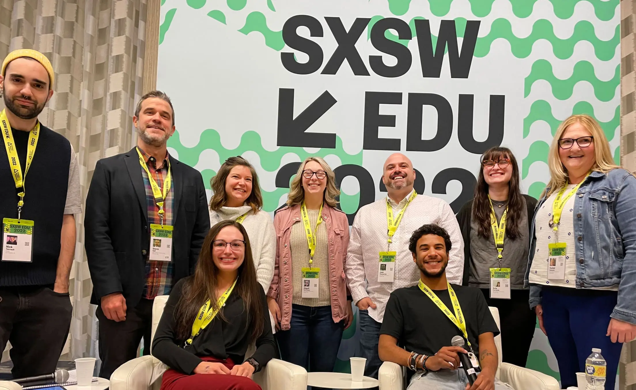 A group of nine people is posing together at the SXSW EDU 2022 event. They are standing and sitting in front of a large, decorative backdrop that reads "SXSW EDU 2022" with a green and white wavy pattern. Each person is wearing a lanyard with an event badge.