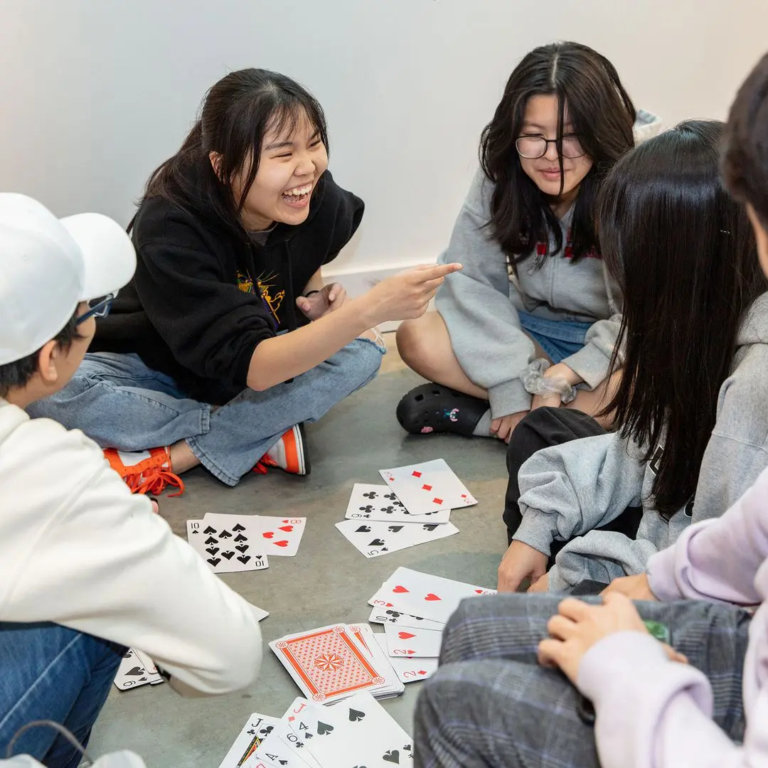 Young people sitting on the floor in a circle, engaging in a lively card game. One girl is laughing, while the others smile and look at the cards.